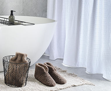 Wit douchegordijn naast een bad met bruine pantoffels en een lichte badmat ervoor