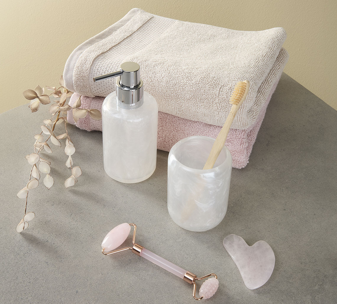 Zandkleurige en roze handdoek naast tandenborstelhouder, zeeppompje en gezichtsroller 