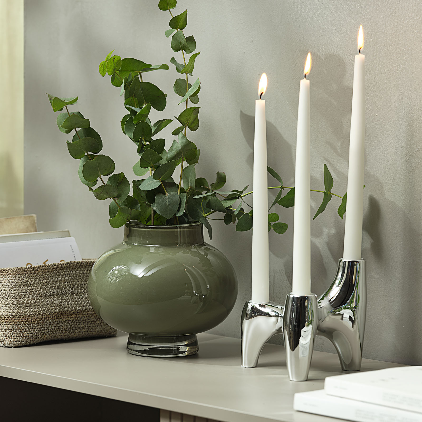 Chromen kandelaar met drie kaarsen ernaast, naast een groene glazen vaas met kunstmatige eucalyptus