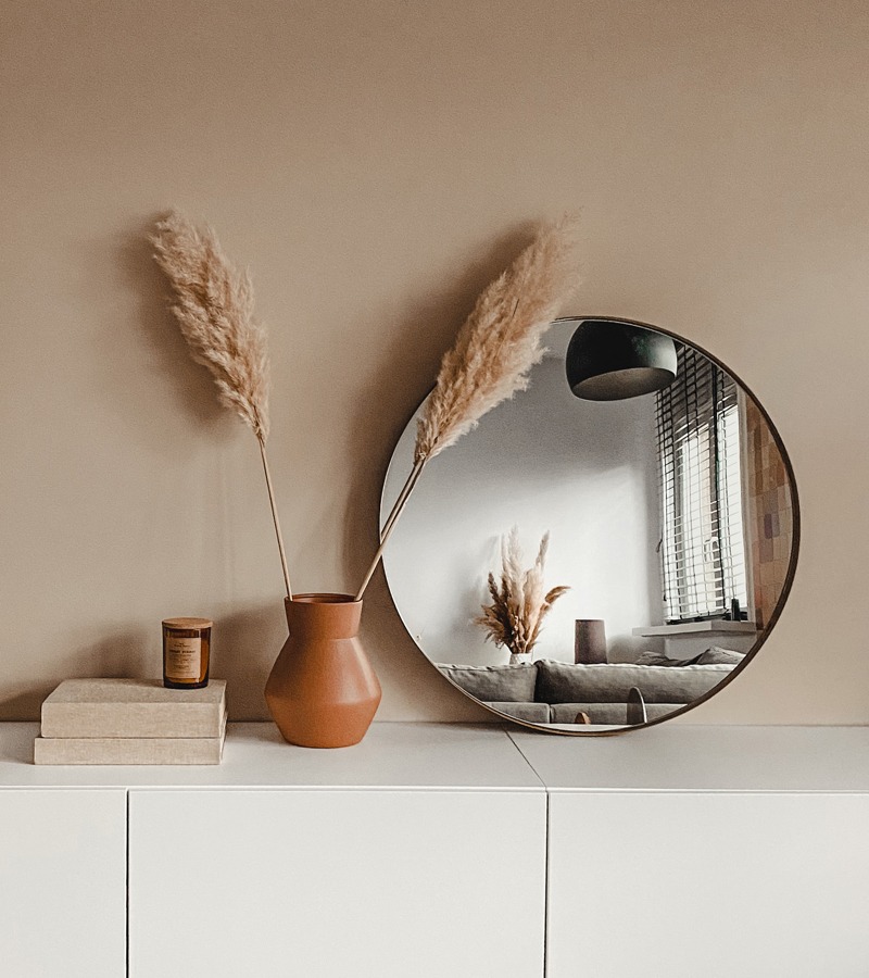 Spiegel MARSTAL goud en vaas SVERRE terracotta Grote ronde spiegel met gouden rand op wit kastje met terracotta vaas met pluimen