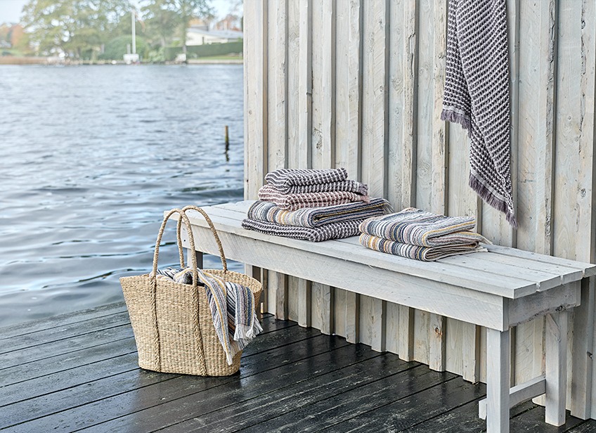 Luxe handdoeken op een bankje en rieten tas bij een meer