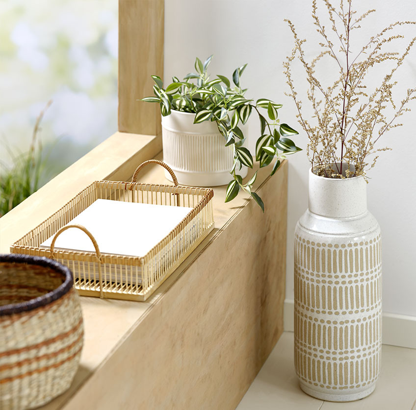 Grote vaas naast vensterbank met bamboe dienblad en witte plantenpot met kunstplant