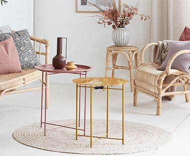 Kleine ronde bijzettafels in oud roze en oker geel op een rond vloerkleed met rotan meubelen in een woonkamer